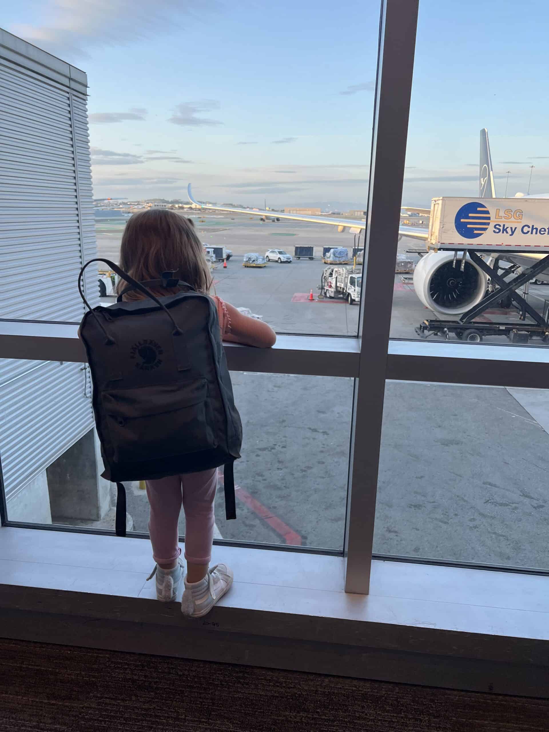 little girl in a backpack looking at planes through an airport window