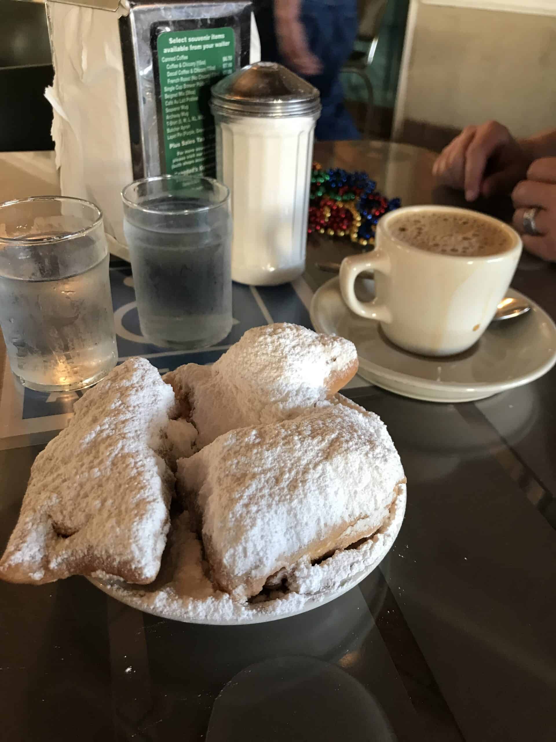 A plate of freshly made beignets dusted with powdered sugar, accompanied by a steaming cup of coffee and glasses of water on a cafe table. The scene captures a casual, inviting dining experience with a touch of Southern charm, ideal for breakfast or a sweet snack.