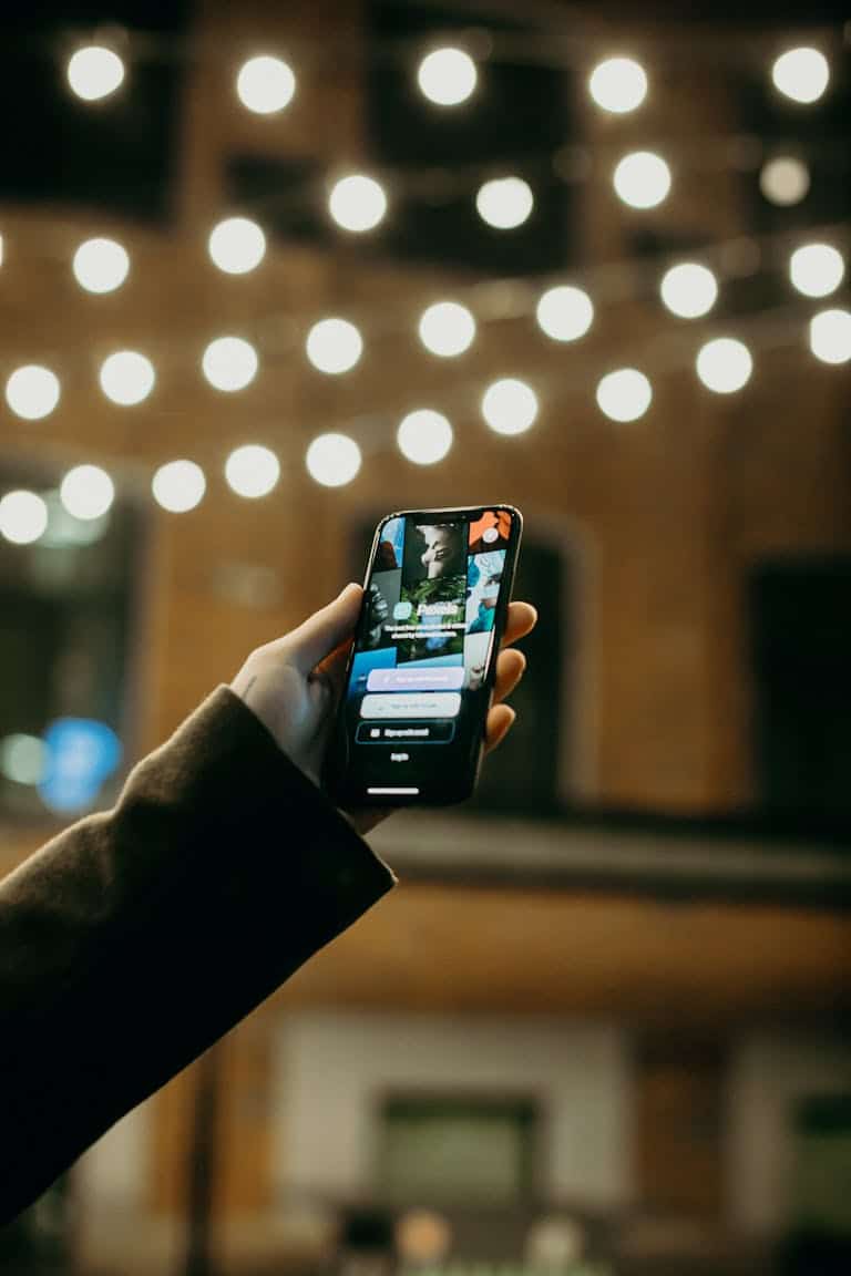 A person holds a smartphone displaying the Wayfaring Four app, designed for travel enthusiasts to discover and document adventures, with warm string lights creating a welcoming atmosphere.