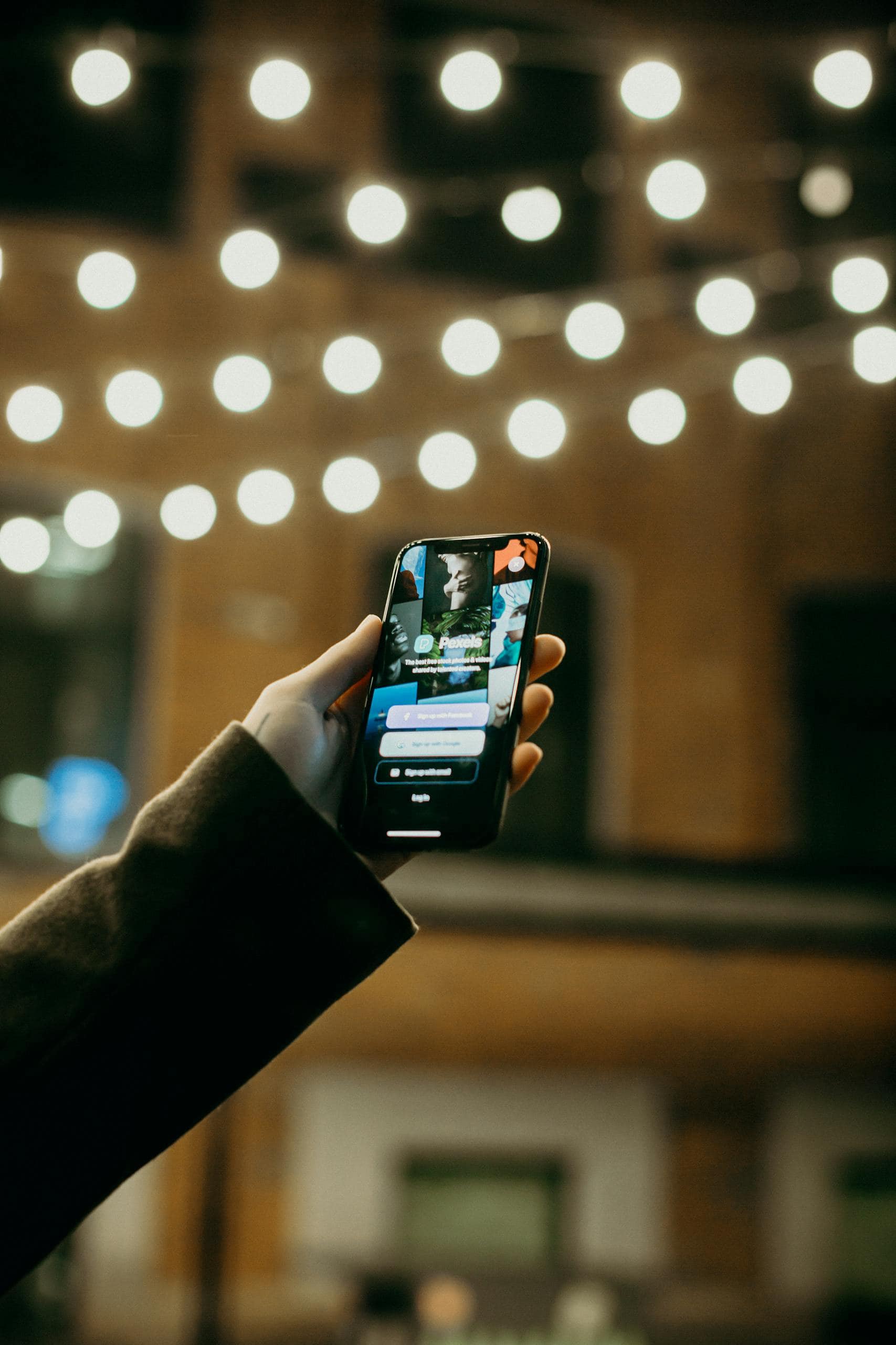 A person holds a smartphone displaying the Wayfaring Four app, designed for travel enthusiasts to discover and document adventures, with warm string lights creating a welcoming atmosphere.