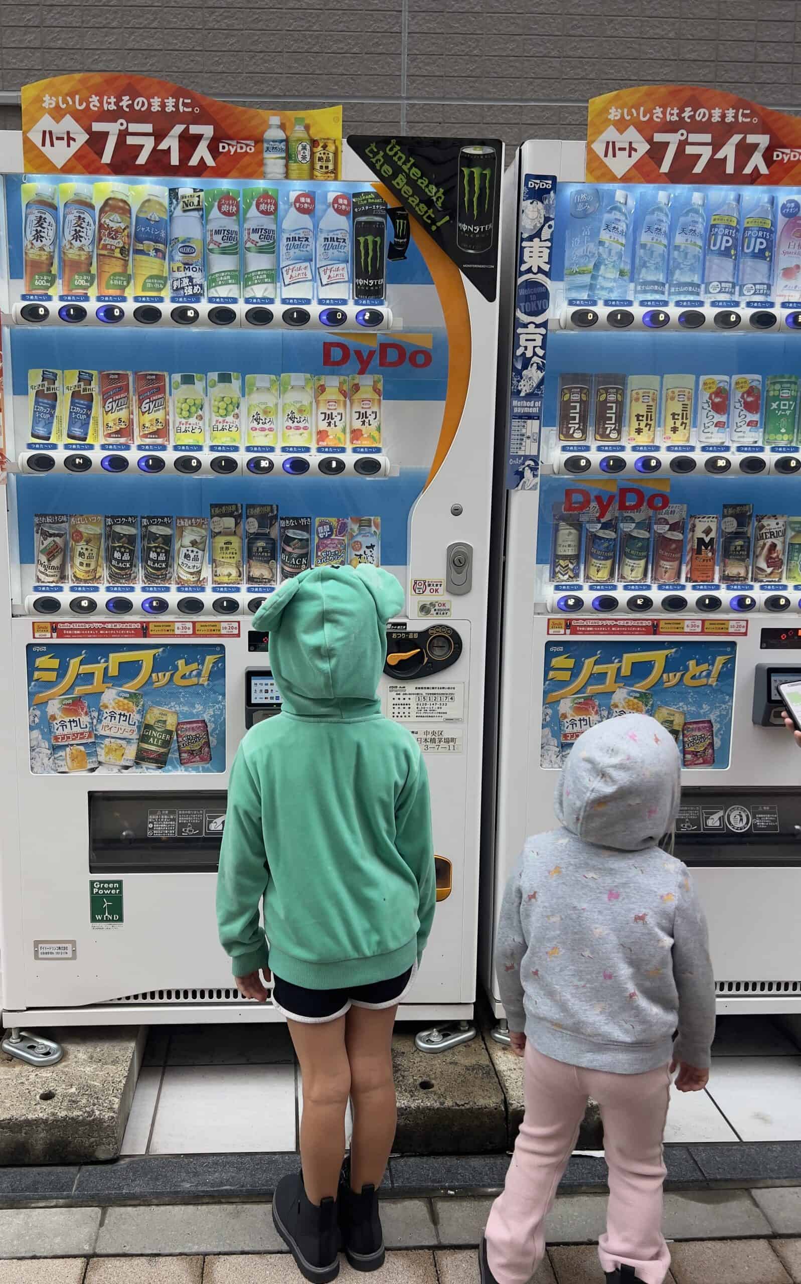 A photo of two children in hoodies standing in front of Japanese vending machines, selecting drinks. The vibrant machines offer a variety of beverages, showcasing Japan's vending machine culture and family-friendly urban scene.