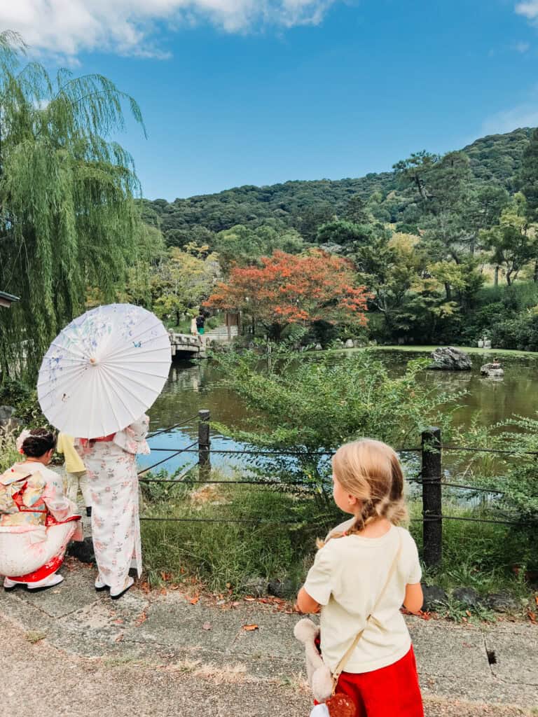 Japanese garden scene with women in kimono, a girl looking at a pond, and colorful autumn foliage.