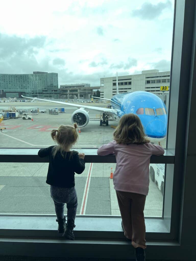 Children looking at airplane through large window at busy airport.