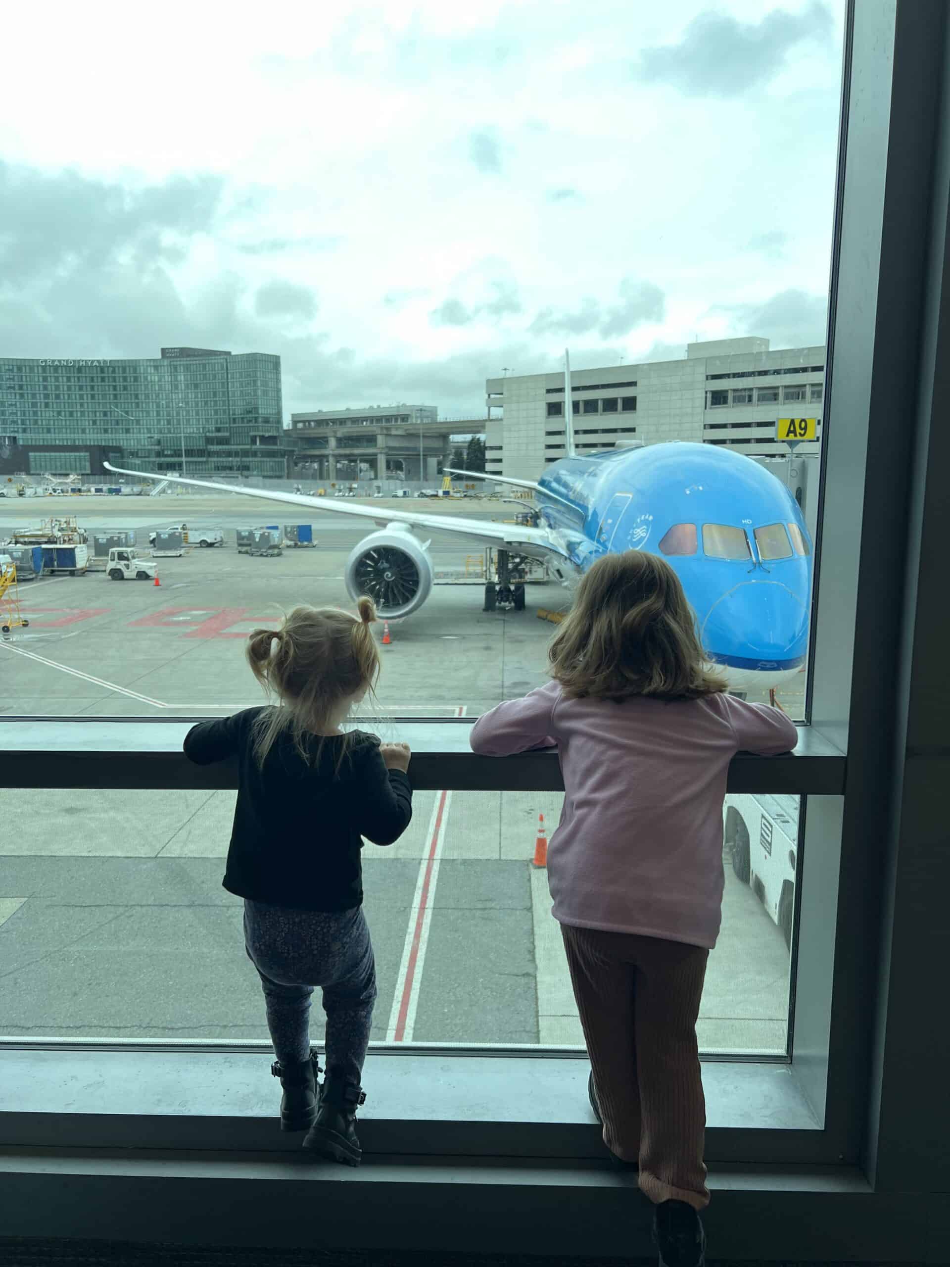 Children looking at airplane through large window at busy airport.